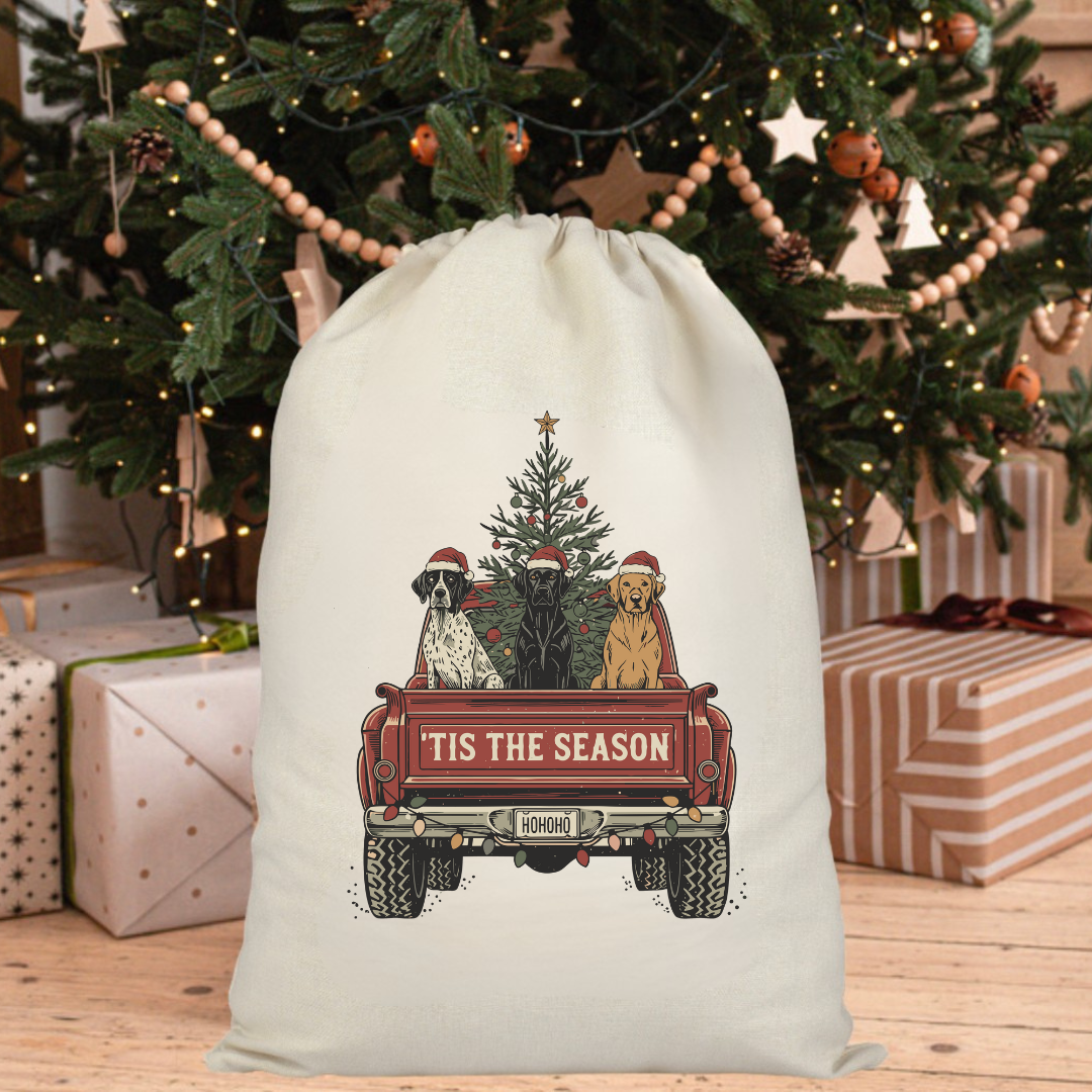 White drawstring bag with Christmas design of dogs in a truck and 'Tis the Season text, placed in front of a decorated Christmas tree and presents.